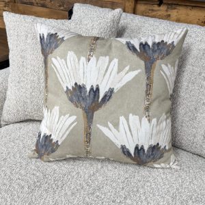 A close-up shot of a stylized botanical accent pillow resting on a light grey textured couch. The square pillow features a repeating pattern of large, artistic white flowers with dark charcoal centers and slender stalks, set against a muted sage green or taupe background. In the soft-focus background, another matching throw pillow is visible alongside a dark wood end table.