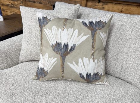 Tactile Neutral Sofa A close-up shot of a stylized botanical accent pillow resting on a light grey textured couch. The square pillow features a repeating pattern of large, artistic white flowers with dark charcoal centers and slender stalks, set against a muted sage green or taupe background. In the soft-focus background, another matching throw pillow is visible alongside a dark wood end table.