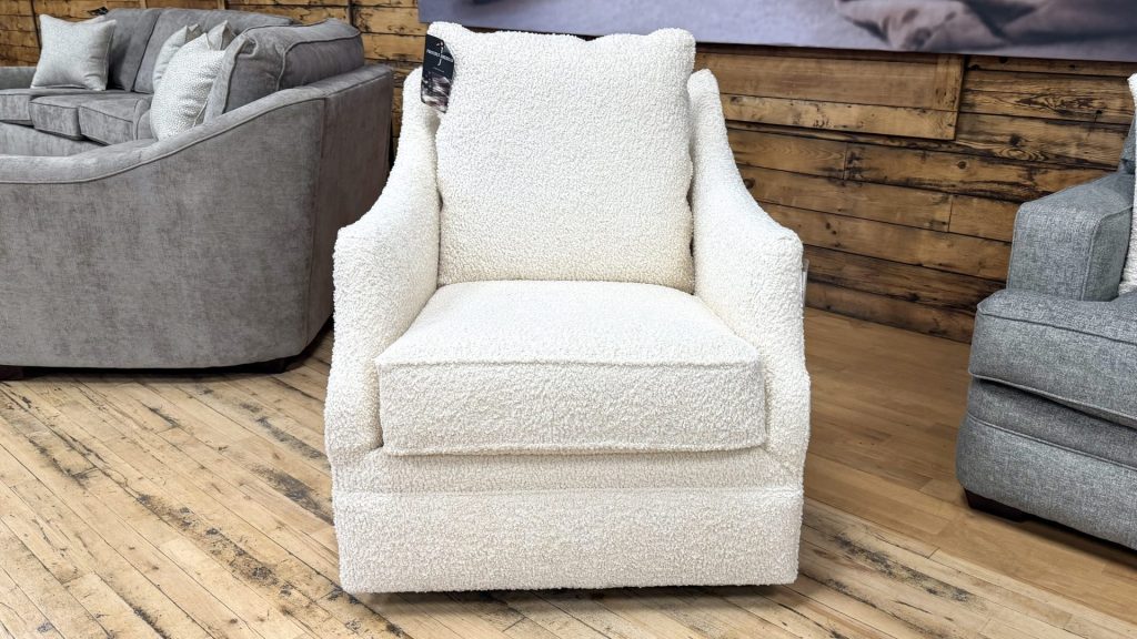 A cream-colored swivel glider accent chair wrapped in high-texture, nubby bouclé fabric stands on a light wood floor in a showroom setting. The chair features a tailored, thick boxed seat cushion, a plush loose back pillow, and elegant, gently sloping track arms. The fully upholstered base conceals the swivel mechanism, extending completely to the floor.