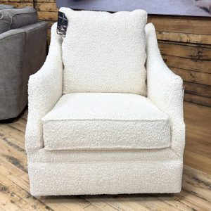 A cream-colored swivel glider accent chair wrapped in high-texture, nubby bouclé fabric stands on a light wood floor in a showroom setting. The chair features a tailored, thick boxed seat cushion, a plush loose back pillow, and elegant, gently sloping track arms. The fully upholstered base conceals the swivel mechanism, extending completely to the floor.
