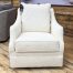 A cream-colored swivel glider accent chair wrapped in high-texture, nubby bouclé fabric stands on a light wood floor in a showroom setting. The chair features a tailored, thick boxed seat cushion, a plush loose back pillow, and elegant, gently sloping track arms. The fully upholstered base conceals the swivel mechanism, extending completely to the floor.