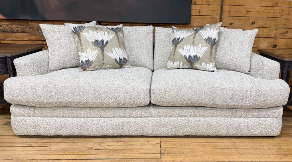 A cozy, transitional-style sofa with light cream and grey textured fabric sits on a light wood floor in front of a rustic wood-planked wall. The sofa features thick, plush seat cushions, low track arms, and a fully upholstered base that goes to the floor. It is styled with two matching textured throw pillows and two accent pillows featuring a large-scale, stylized white and grey floral pattern.