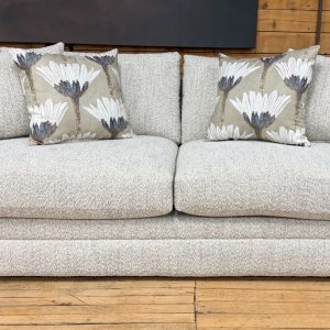 A cozy, transitional-style sofa with light cream and grey textured fabric sits on a light wood floor in front of a rustic wood-planked wall. The sofa features thick, plush seat cushions, low track arms, and a fully upholstered base that goes to the floor. It is styled with two matching textured throw pillows and two accent pillows featuring a large-scale, stylized white and grey floral pattern.