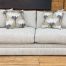 A cozy, transitional-style sofa with light cream and grey textured fabric sits on a light wood floor in front of a rustic wood-planked wall. The sofa features thick, plush seat cushions, low track arms, and a fully upholstered base that goes to the floor. It is styled with two matching textured throw pillows and two accent pillows featuring a large-scale, stylized white and grey floral pattern.