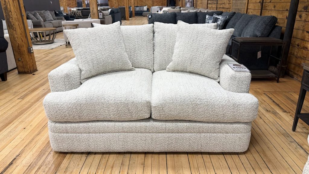 A cozy, transitional-style loveseat wrapped in light cream and grey textured bouclé fabric sits on a light hardwood floor in a furniture showroom. The two-seater couch features thick, plush seat cushions, low track arms, a fully upholstered base, and matching textured throw pillows. In the background, other sofas and rustic wooden pillars are visible across the showroom floor.