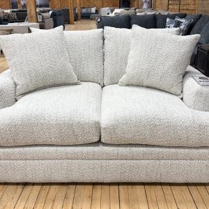 A cozy, transitional-style loveseat wrapped in light cream and grey textured bouclé fabric sits on a light hardwood floor in a furniture showroom. The two-seater couch features thick, plush seat cushions, low track arms, a fully upholstered base, and matching textured throw pillows. In the background, other sofas and rustic wooden pillars are visible across the showroom floor.