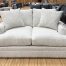 A cozy, transitional-style loveseat wrapped in light cream and grey textured bouclé fabric sits on a light hardwood floor in a furniture showroom. The two-seater couch features thick, plush seat cushions, low track arms, a fully upholstered base, and matching textured throw pillows. In the background, other sofas and rustic wooden pillars are visible across the showroom floor.
