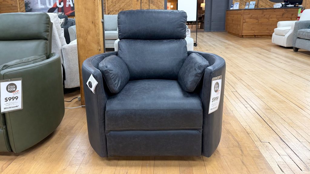 Modern charcoal grey faux-leather recliner chair with built-in side bolster pillows, displayed on a light hardwood showroom floor at The Stock Room.