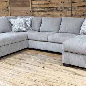 A large, U-shaped sectional sofa wrapped in a light grey, subtly textured woven fabric sits on a light hardwood floor in a showroom. The sectional features a left-arm-facing chaise lounge, a central seating area, and a right-arm-facing chaise lounge, all supported by short, dark wood block feet. It is styled with multiple matching back cushions and four decorative accent pillows featuring geometric and abstract patterns in neutral, cream, and terracotta tones. A rustic wood-planked wall serves as the backdrop.
