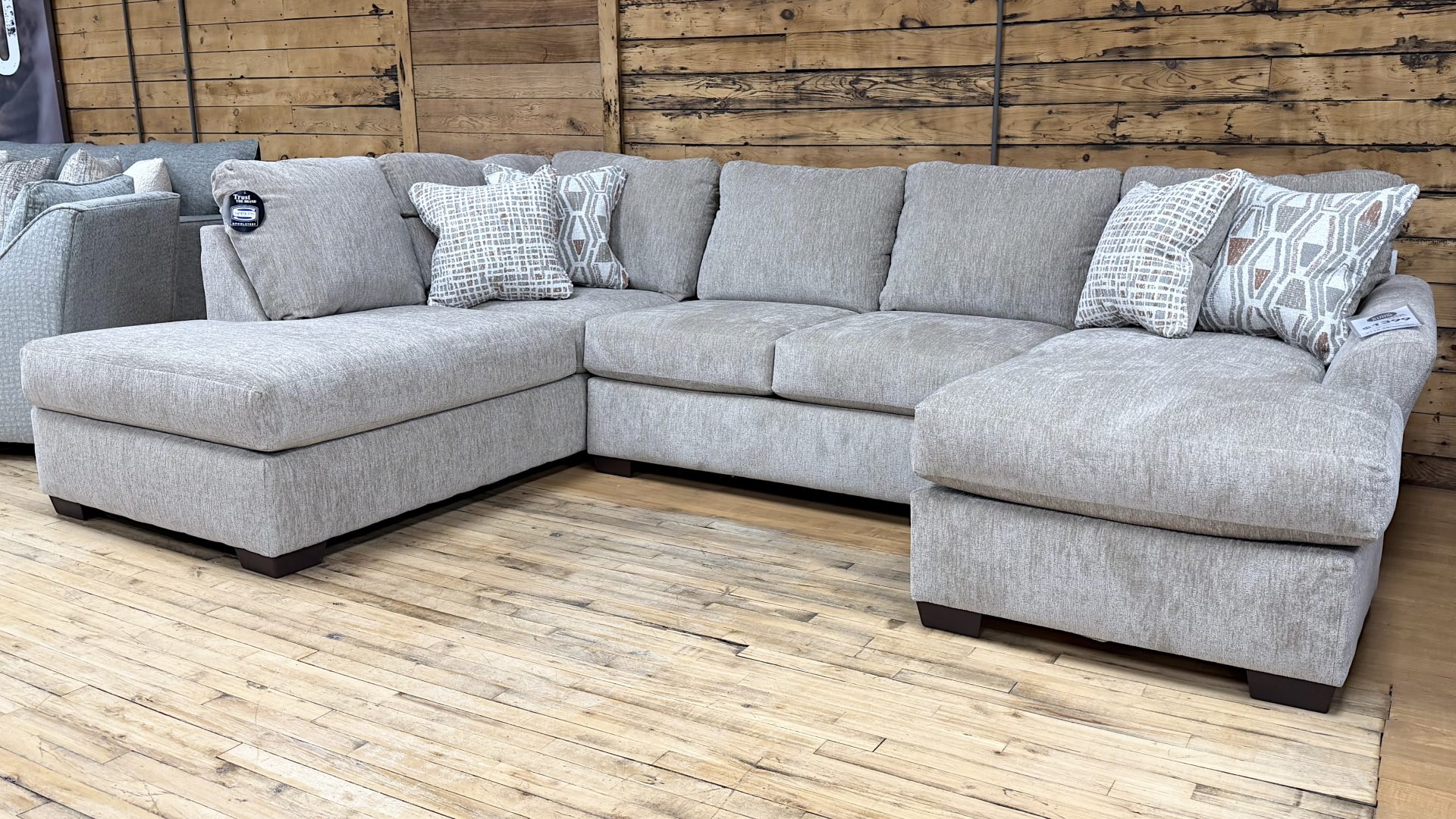 Tezza-4648 A large, U-shaped sectional sofa wrapped in a light grey, subtly textured woven fabric sits on a light hardwood floor in a showroom. The sectional features a left-arm-facing chaise lounge, a central seating area, and a right-arm-facing chaise lounge, all supported by short, dark wood block feet. It is styled with multiple matching back cushions and four decorative accent pillows featuring geometric and abstract patterns in neutral, cream, and terracotta tones. A rustic wood-planked wall serves as the backdrop.
