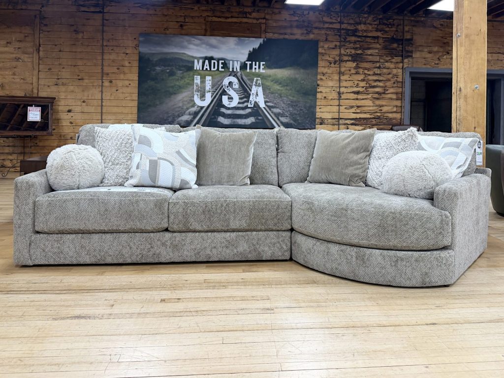 Large beige textured tweed sectional sofa featuring a rounded cuddler chaise on the right side, styled with fuzzy round pillows and patterned throw cushions on a hardwood showroom floor.