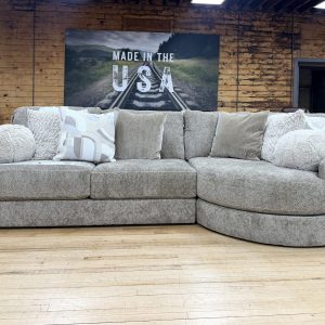 Large beige textured tweed sectional sofa featuring a rounded cuddler chaise on the right side, styled with fuzzy round pillows and patterned throw cushions on a hardwood showroom floor.