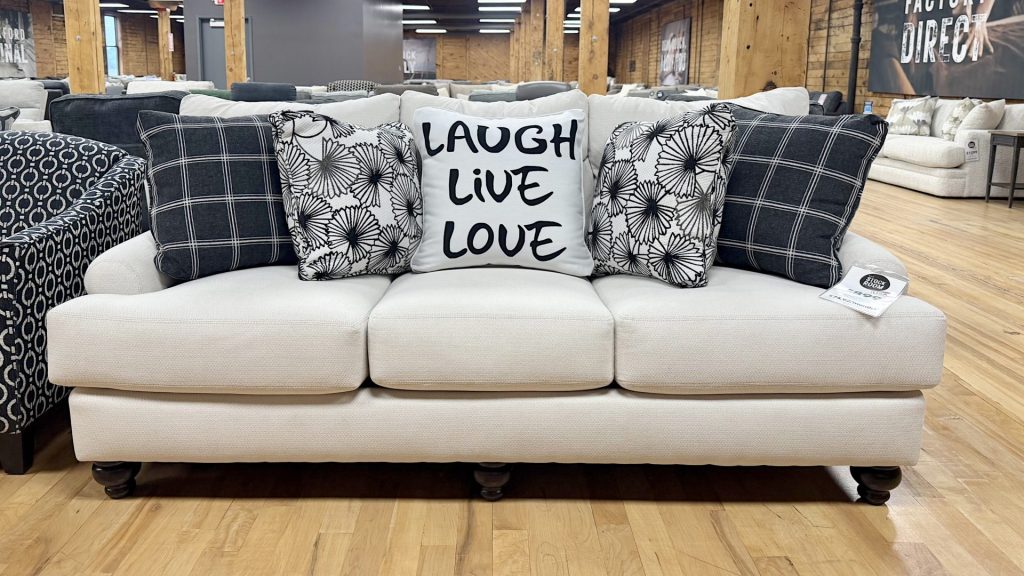 Cream-colored upholstered transitional sofa featuring dark turned wood bun feet and five decorative accent pillows, including black plaid patterns and a center pillow reading "LAUGH LIVE LOVE" on a hardwood showroom floor.