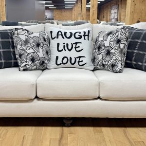 Cream-colored upholstered transitional sofa featuring dark turned wood bun feet and five decorative accent pillows, including black plaid patterns and a center pillow reading "LAUGH LIVE LOVE" on a hardwood showroom floor.