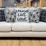 Cream-colored upholstered transitional sofa featuring dark turned wood bun feet and five decorative accent pillows, including black plaid patterns and a center pillow reading "LAUGH LIVE LOVE" on a hardwood showroom floor.