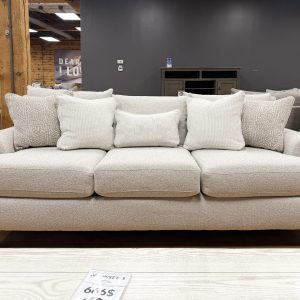 Plush three-cushion sofa upholstered in a textured cream heathered fabric, featuring soft rolled arms, dark tapered wooden legs, and styled with five neutral accent pillows on a showroom hardwood floor.