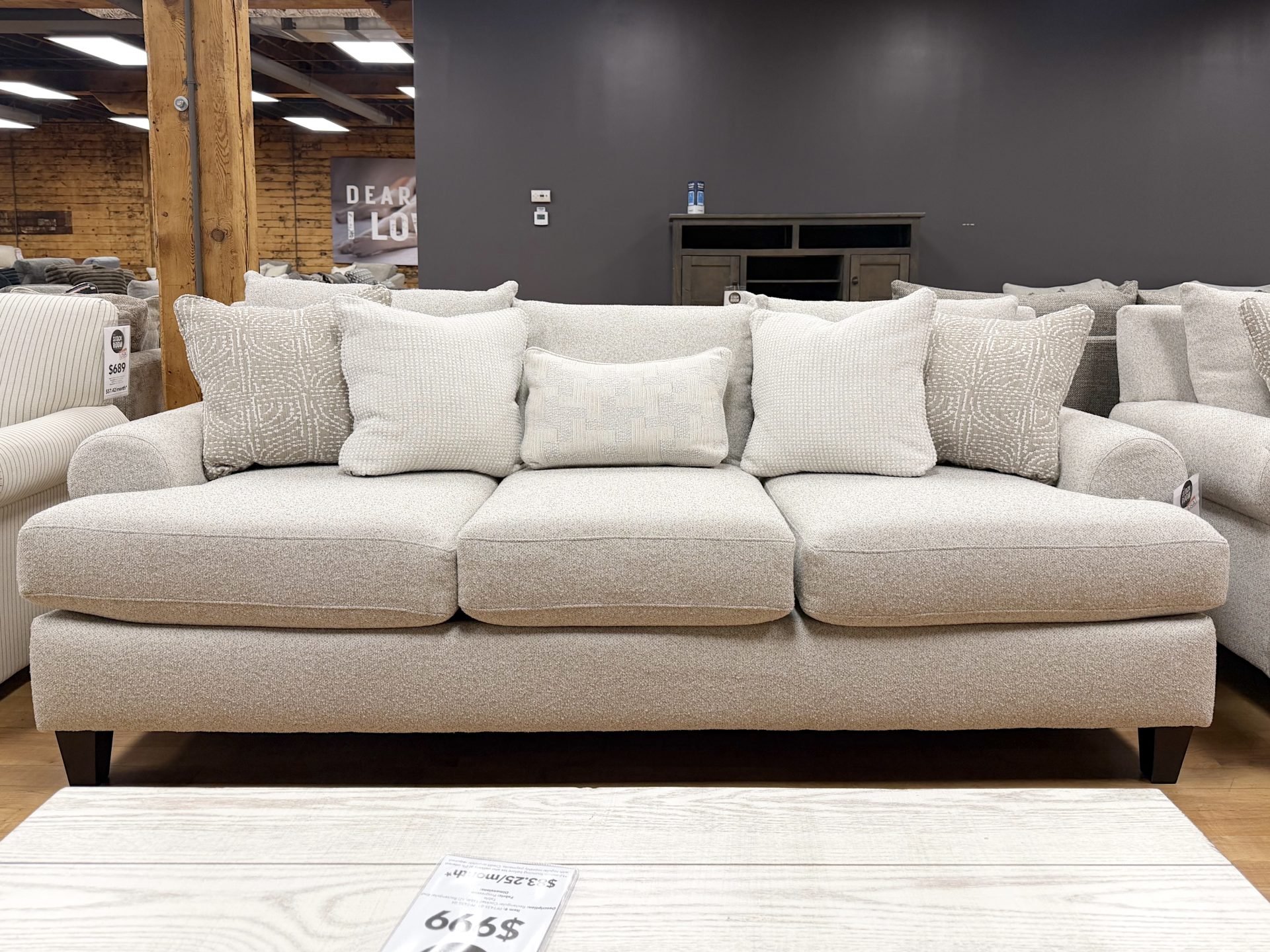 Tezza-7476 Plush three-cushion sofa upholstered in a textured cream heathered fabric, featuring soft rolled arms, dark tapered wooden legs, and styled with five neutral accent pillows on a showroom hardwood floor.