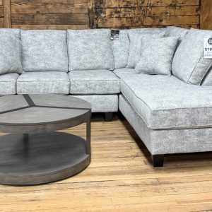 Light gray textured sectional sofa with a right-side chaise, shown in a showroom with wooden walls and a round two-tier coffee table.