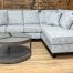 Light gray textured sectional sofa with a right-side chaise, shown in a showroom with wooden walls and a round two-tier coffee table.