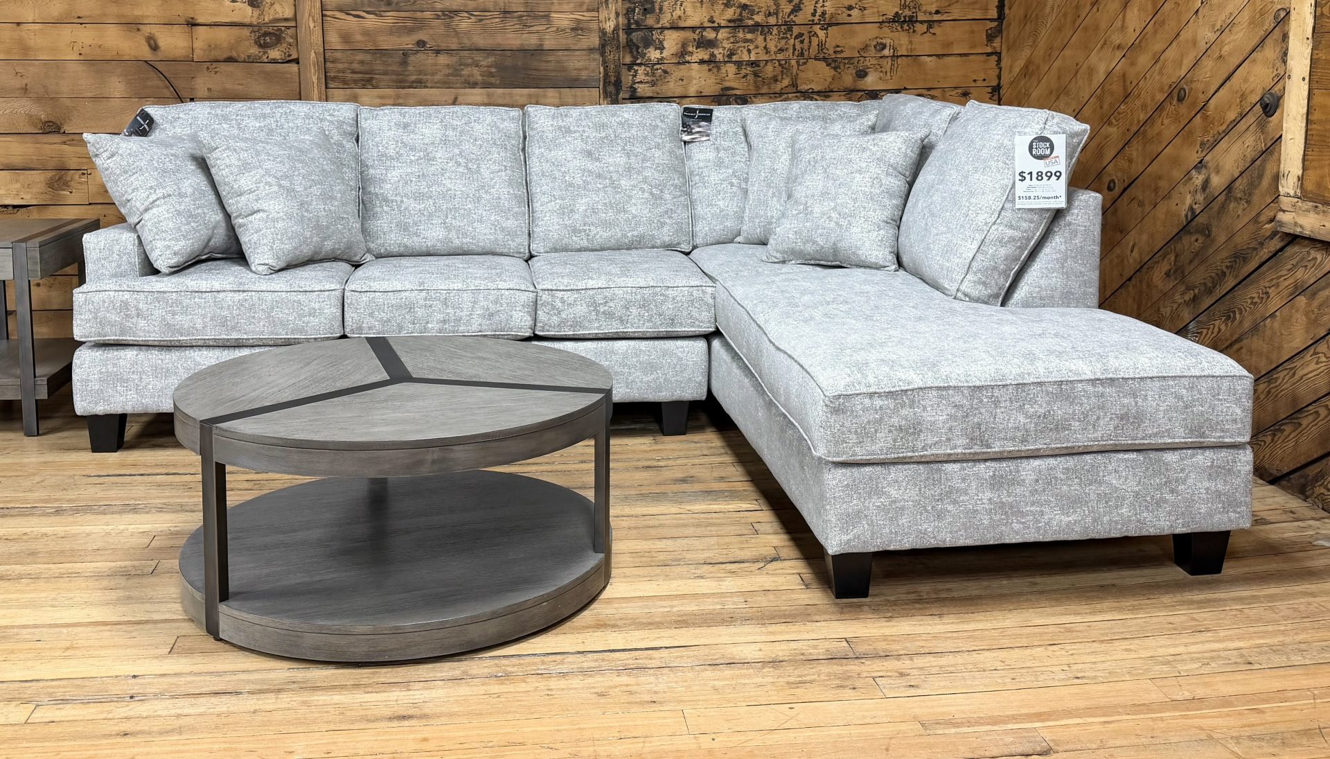 875E8410-59F2-40F6-BB5F-7362E5636EF5 Light gray textured sectional sofa with a right-side chaise, shown in a showroom with wooden walls and a round two-tier coffee table.