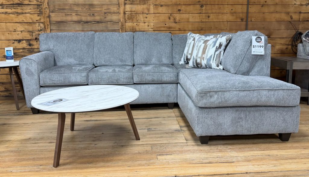 Light gray textured sectional sofa with a right-side chaise, accessorized with patterned throw pillows, positioned in a showroom setting with a white marble-top coffee table.