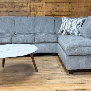 Light gray textured sectional sofa with a right-side chaise, accessorized with patterned throw pillows, positioned in a showroom setting with a white marble-top coffee table.