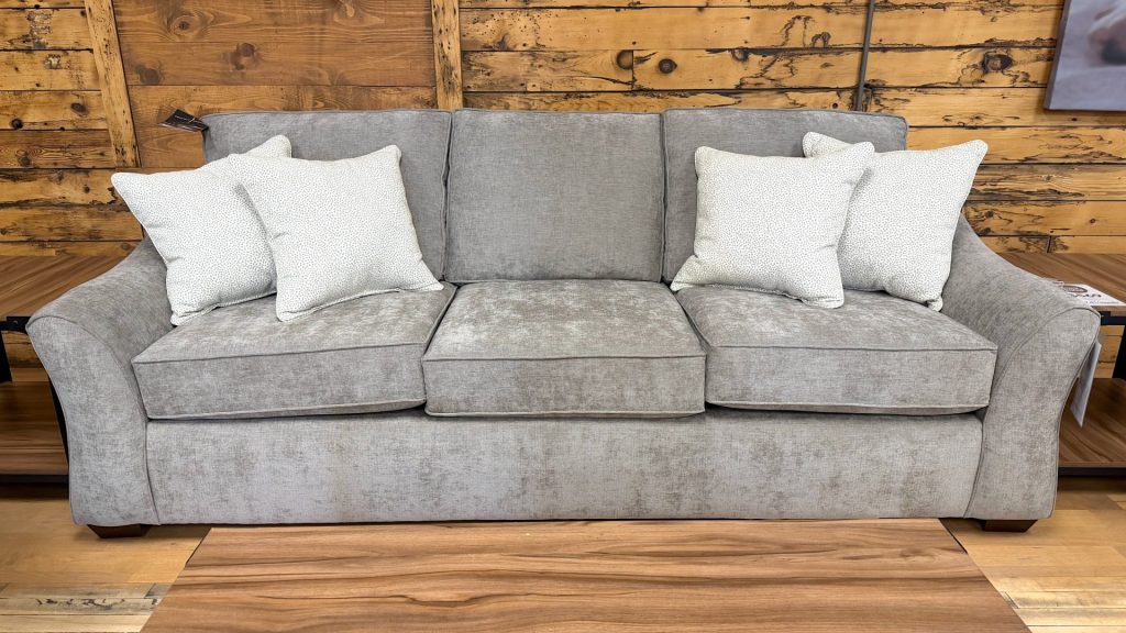 Plush three-cushion sofa upholstered in a textured heathered taupe fabric, featuring wide flared arms, dark wooden feet, and styled with four cream and grey dotted accent pillows on a showroom hardwood floor.