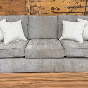 Plush three-cushion sofa upholstered in a textured heathered taupe fabric, featuring wide flared arms, dark wooden feet, and styled with four cream and grey dotted accent pillows on a showroom hardwood floor.