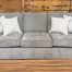 Plush three-cushion sofa upholstered in a textured heathered taupe fabric, featuring wide flared arms, dark wooden feet, and styled with four cream and grey dotted accent pillows on a showroom hardwood floor.