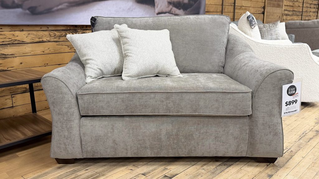 Plush oversized chair and a half upholstered in a textured heathered taupe fabric, featuring wide flared arms, dark wooden block feet, and styled with two cream and grey dotted accent pillows on a showroom hardwood floor.
