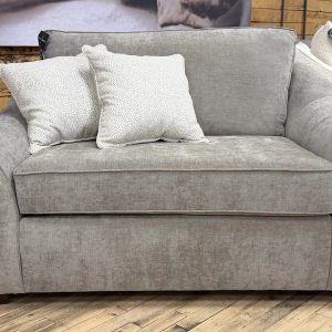 Plush oversized chair and a half upholstered in a textured heathered taupe fabric, featuring wide flared arms, dark wooden block feet, and styled with two cream and grey dotted accent pillows on a showroom hardwood floor.