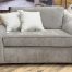 Plush oversized chair and a half upholstered in a textured heathered taupe fabric, featuring wide flared arms, dark wooden block feet, and styled with two cream and grey dotted accent pillows on a showroom hardwood floor.