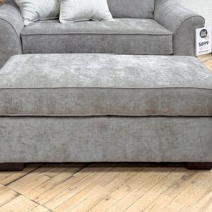 Large rectangular ottoman upholstered in a textured heathered taupe fabric, featuring a thick box cushion top and dark wooden block feet, positioned on a showroom hardwood floor.