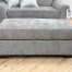 Large rectangular ottoman upholstered in a textured heathered taupe fabric, featuring a thick box cushion top and dark wooden block feet, positioned on a showroom hardwood floor.