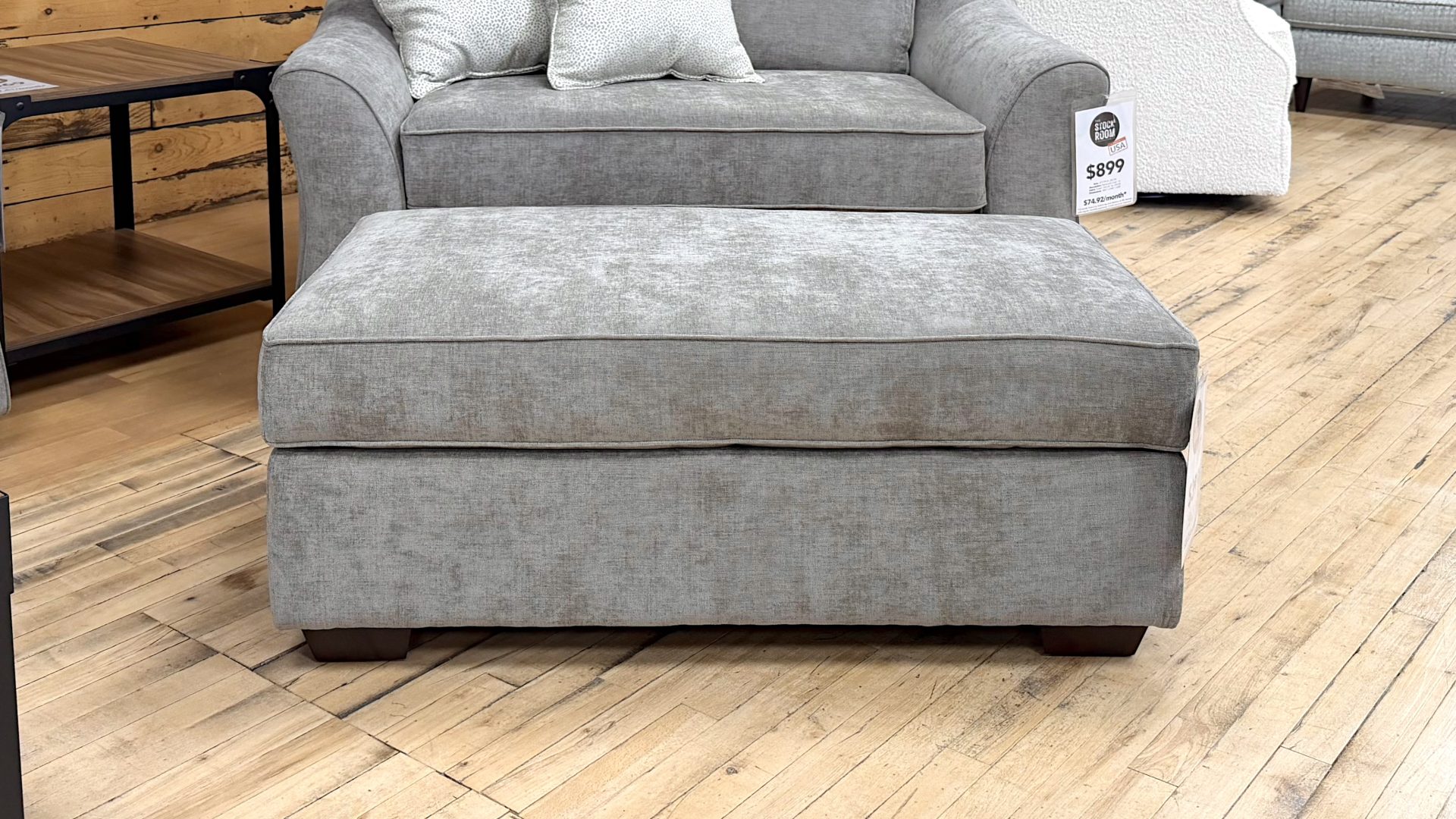 Tezza-9757 Large rectangular ottoman upholstered in a textured heathered taupe fabric, featuring a thick box cushion top and dark wooden block feet, positioned on a showroom hardwood floor.